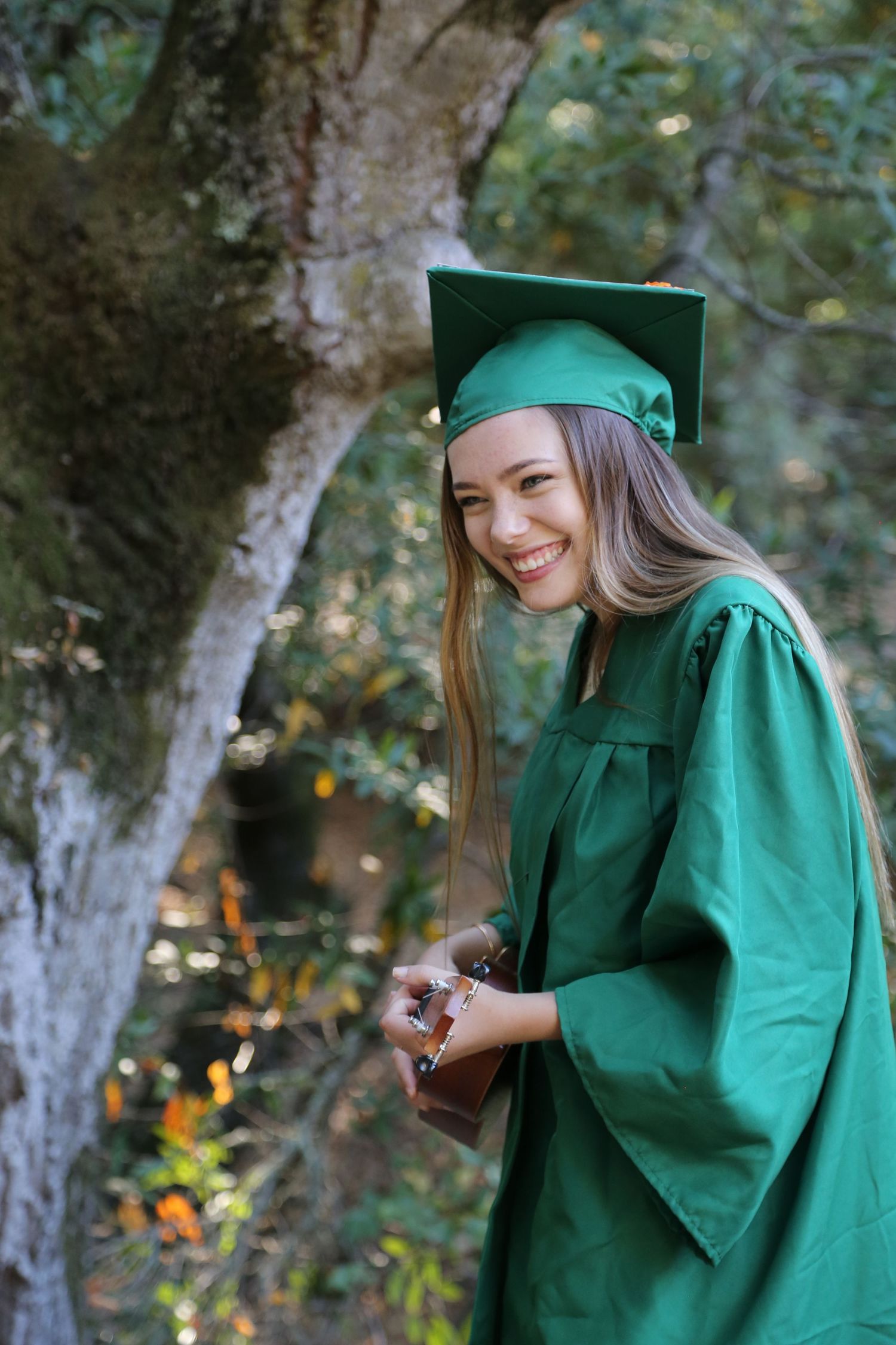 Smiling graduate in a green cap and gown holds a small item beside a mossy oak tree surrounded by autumn foliage.