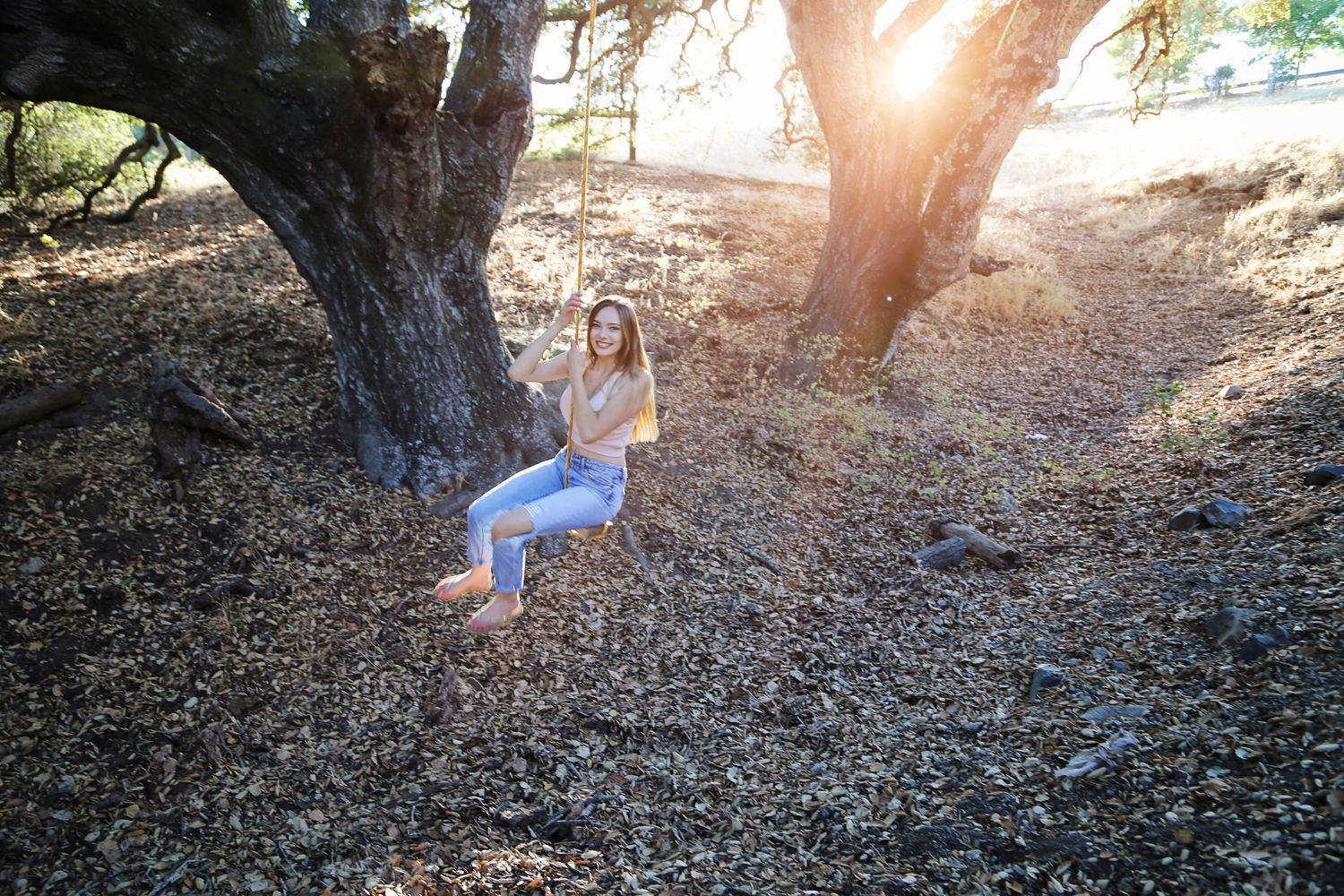 Young woman in jeans sits on a rope swing hanging from an oak tree in a sunlit, golden-lit outdoor setting.
