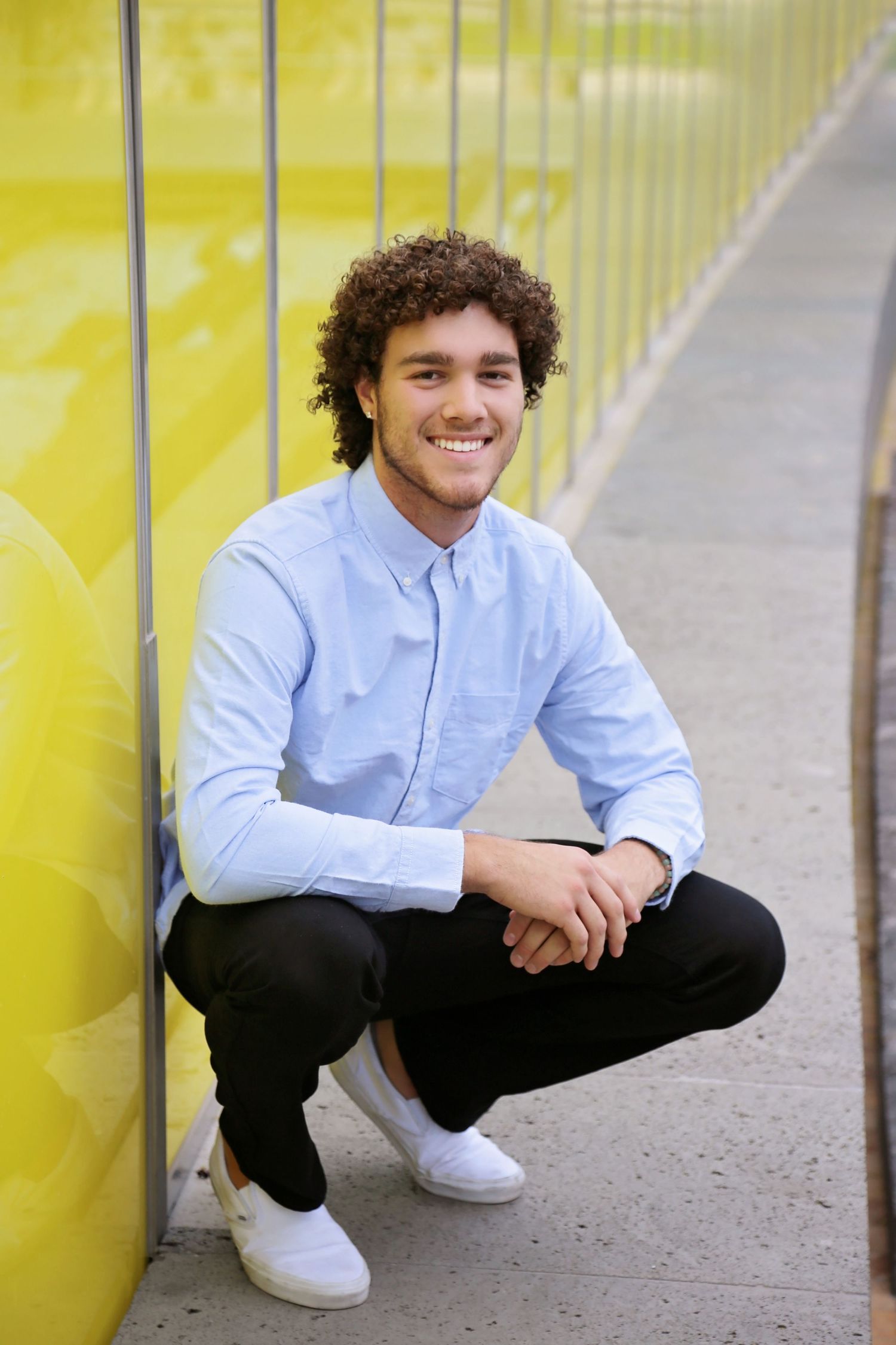 Smiling young man with curly hair crouches in a light blue shirt and white sneakers beside a bright yellow wall.