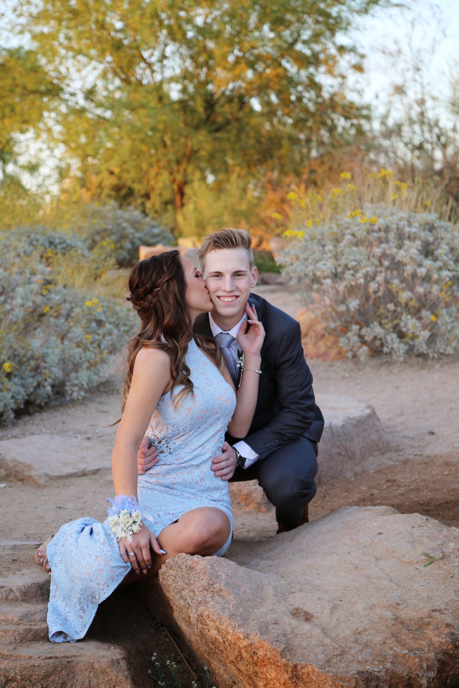 Young couple in formal attire share a kiss on the cheek while seated on rocks in a desert garden, the woman wearing a corsage.