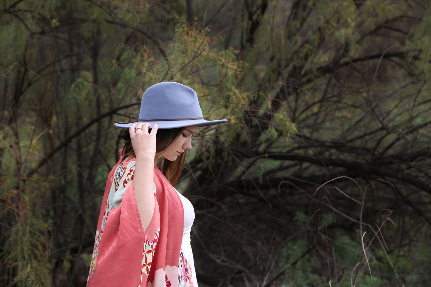 Young woman in a gray hat and coral floral kimono stands in profile among tree branches, holding the hat brim.
