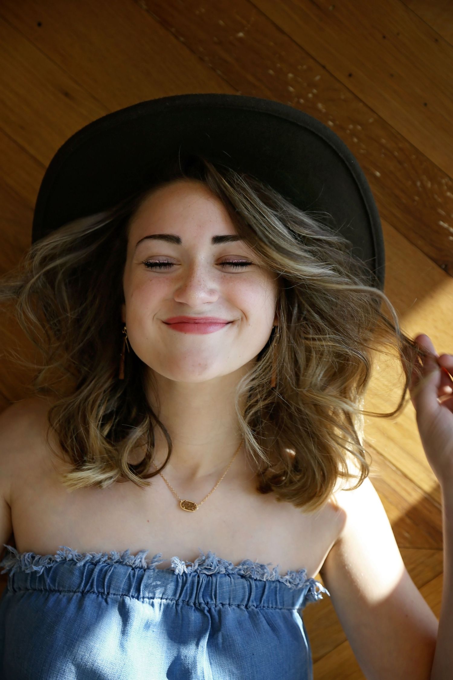 Smiling young woman with closed eyes lies on a wooden floor wearing a black wide-brim hat and a denim off-the-shoulder top.