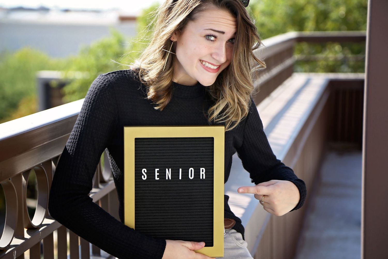Young woman in a black long-sleeve top smiles on a balcony holding a letter board that reads SENIOR for senior portraits.