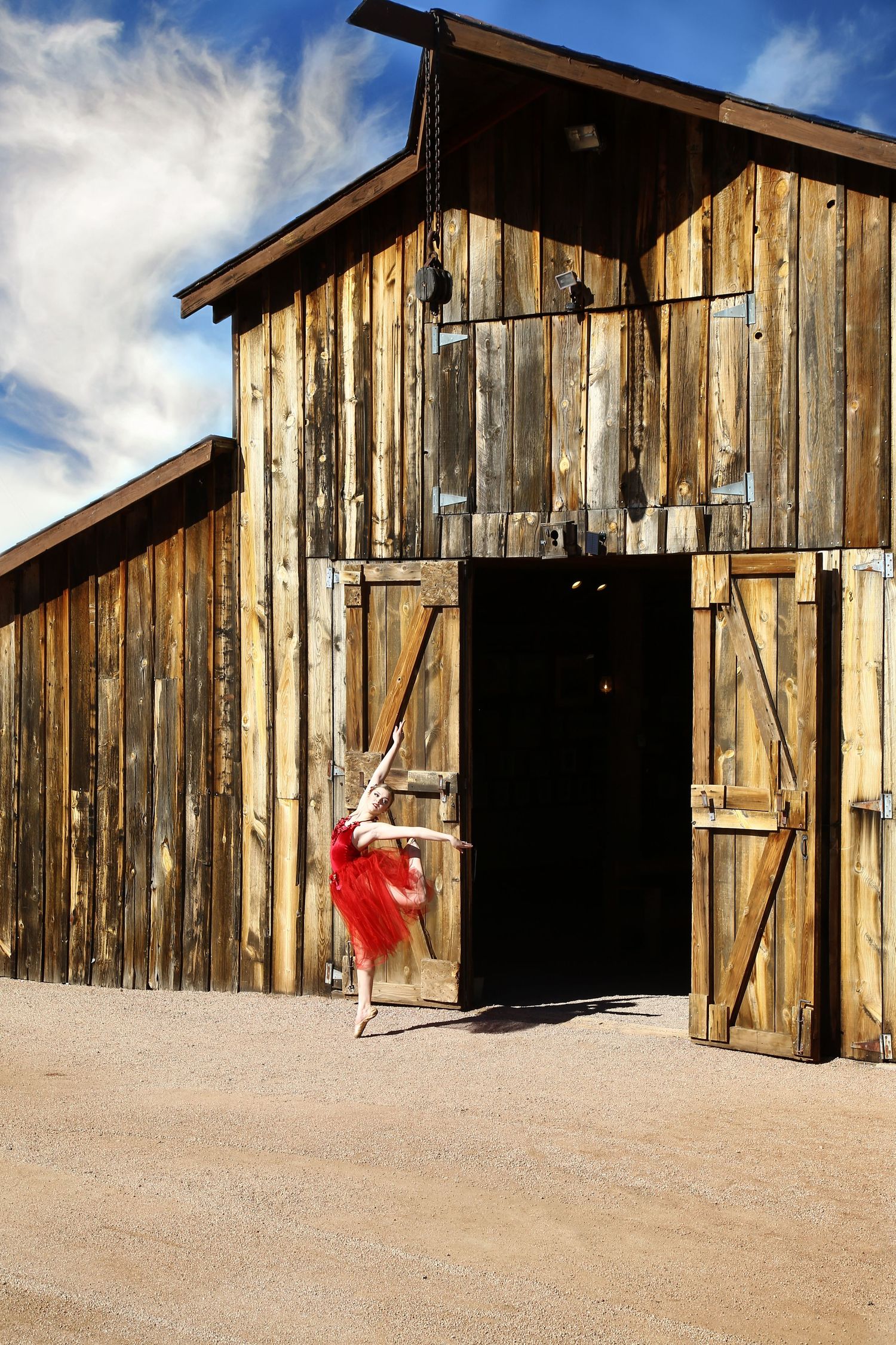 Ballerina in a red tutu poses in an arabesque in front of a rustic wooden barn with open doors under a bright blue sky.