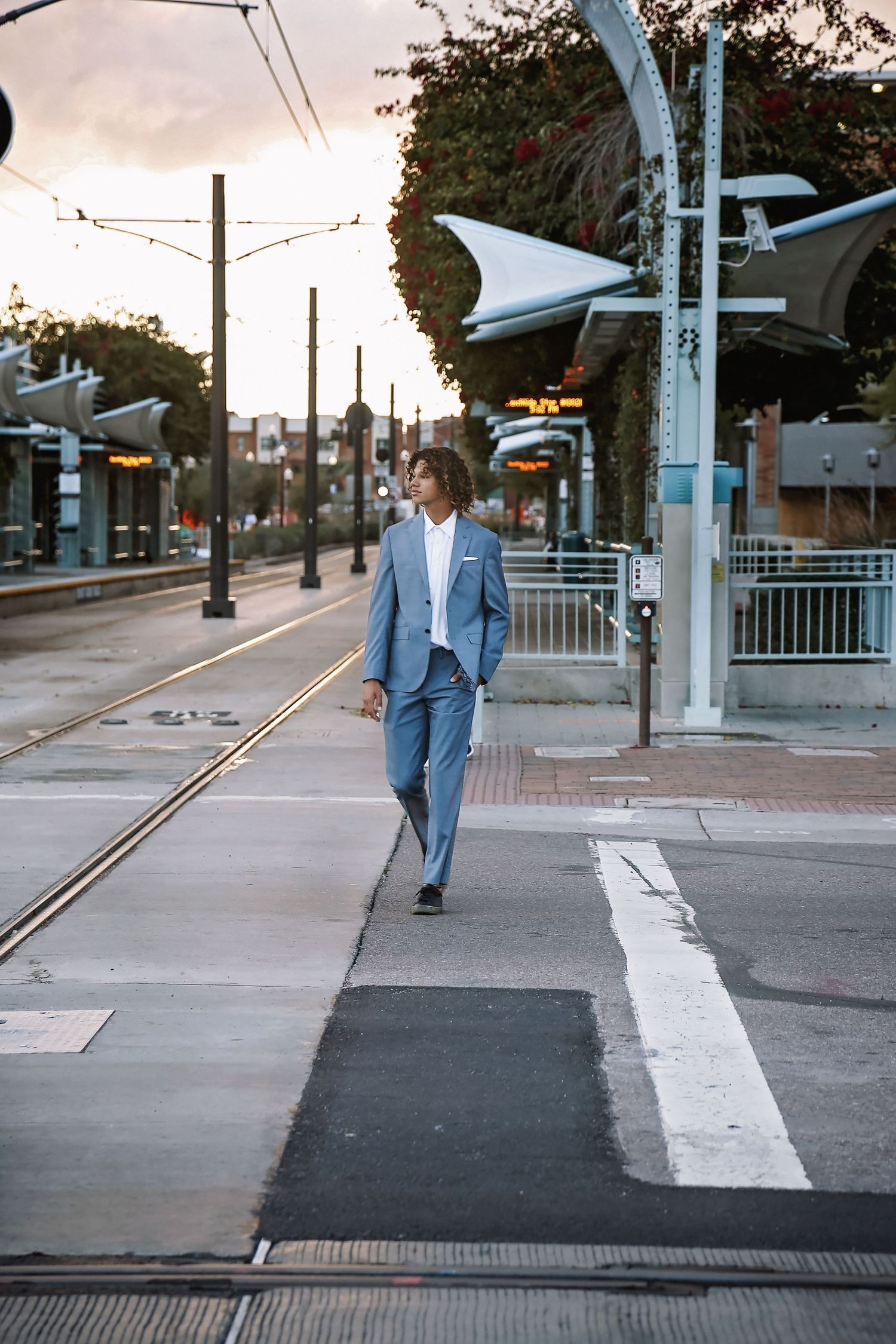 Young man in a light blue suit walks across light rail tracks at a city station during sunset for a senior portrait.