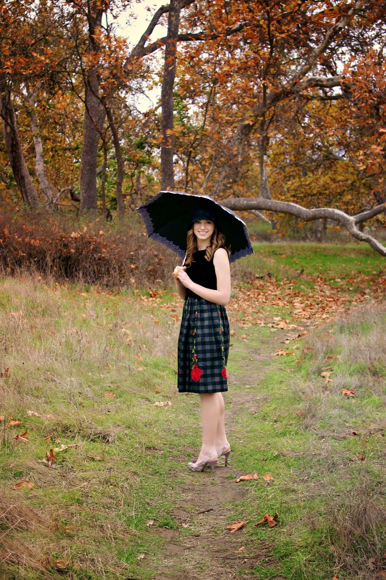 Young woman in a plaid skirt and black top holds a black umbrella on a grassy path surrounded by autumn trees and fallen leaves.