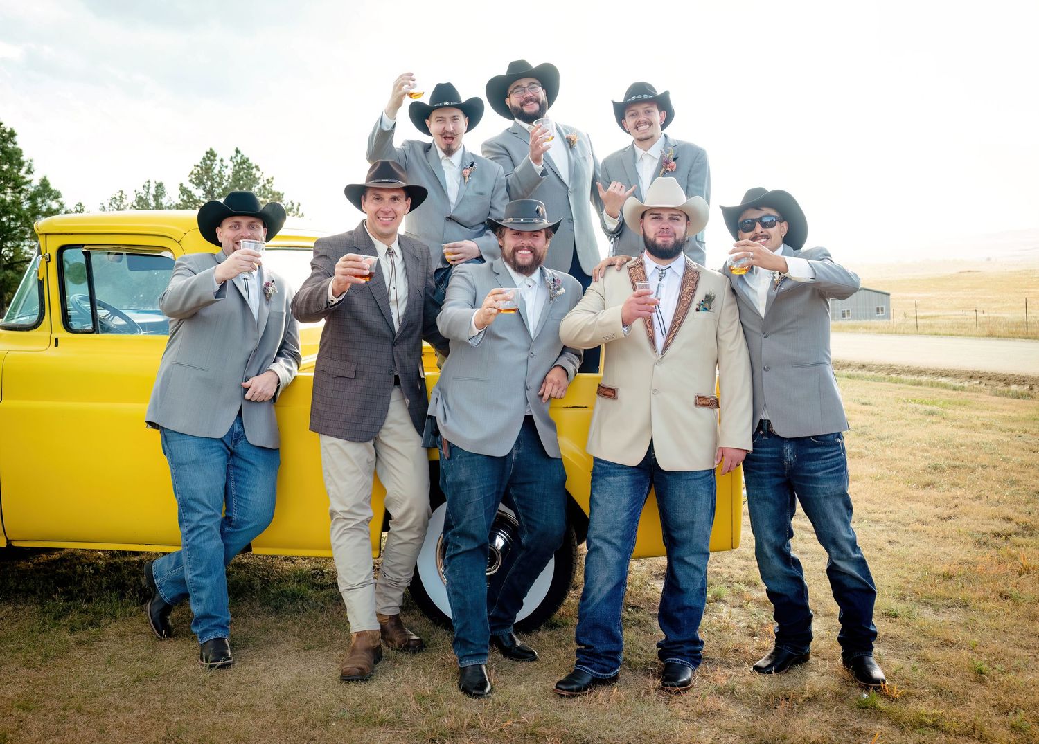 Group of groomsmen in matching blazers and cowboy hats posing beside a vintage yellow truck at an outdoor wedding.
