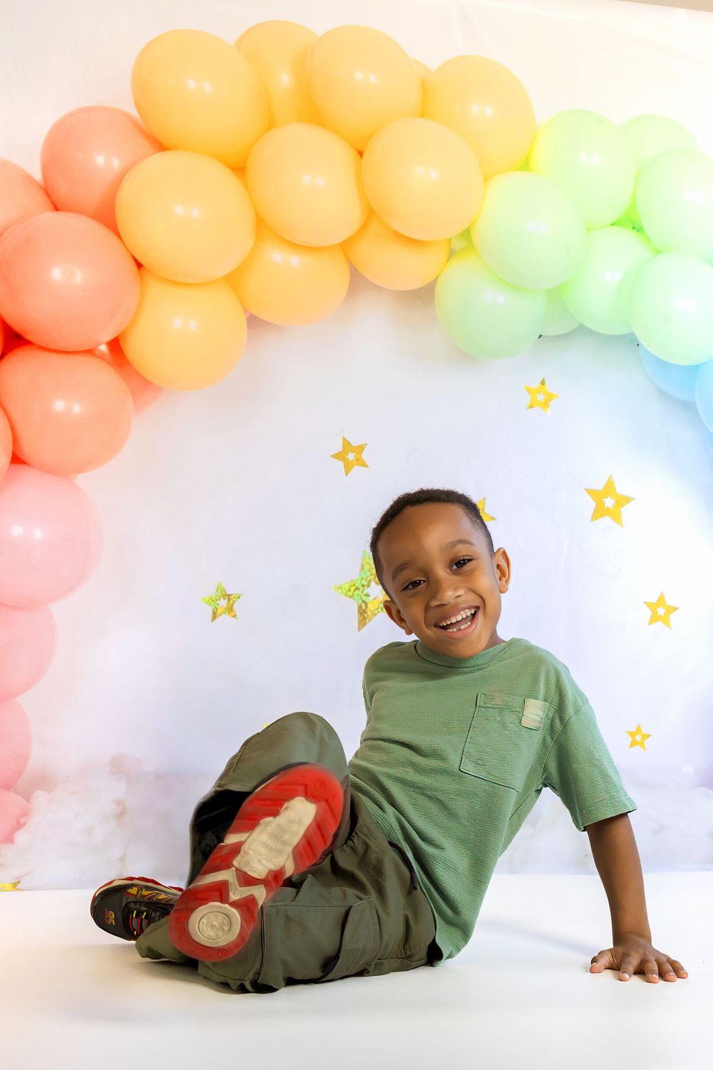 A smiling young boy sits on a white floor in front of a colorful rainbow balloon arch with gold stars.