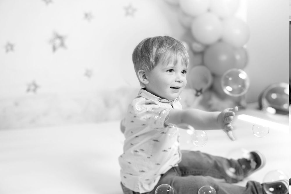 Black and white photo of a toddler boy reaching for soap bubbles at a birthday party with balloon decorations.