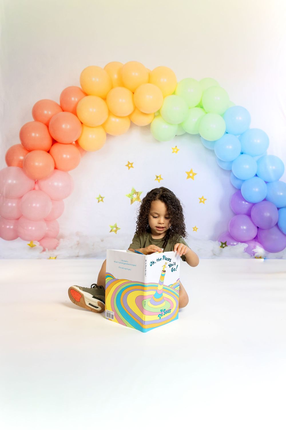 Child reading a colorful book seated before a rainbow balloon arch with gold stars on white backdrop.