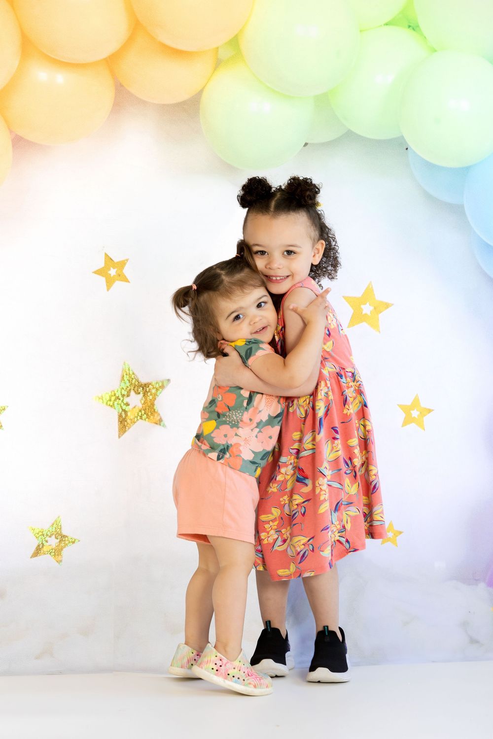 Two young girls hugging and smiling in front of a colorful balloon arch with gold stars.