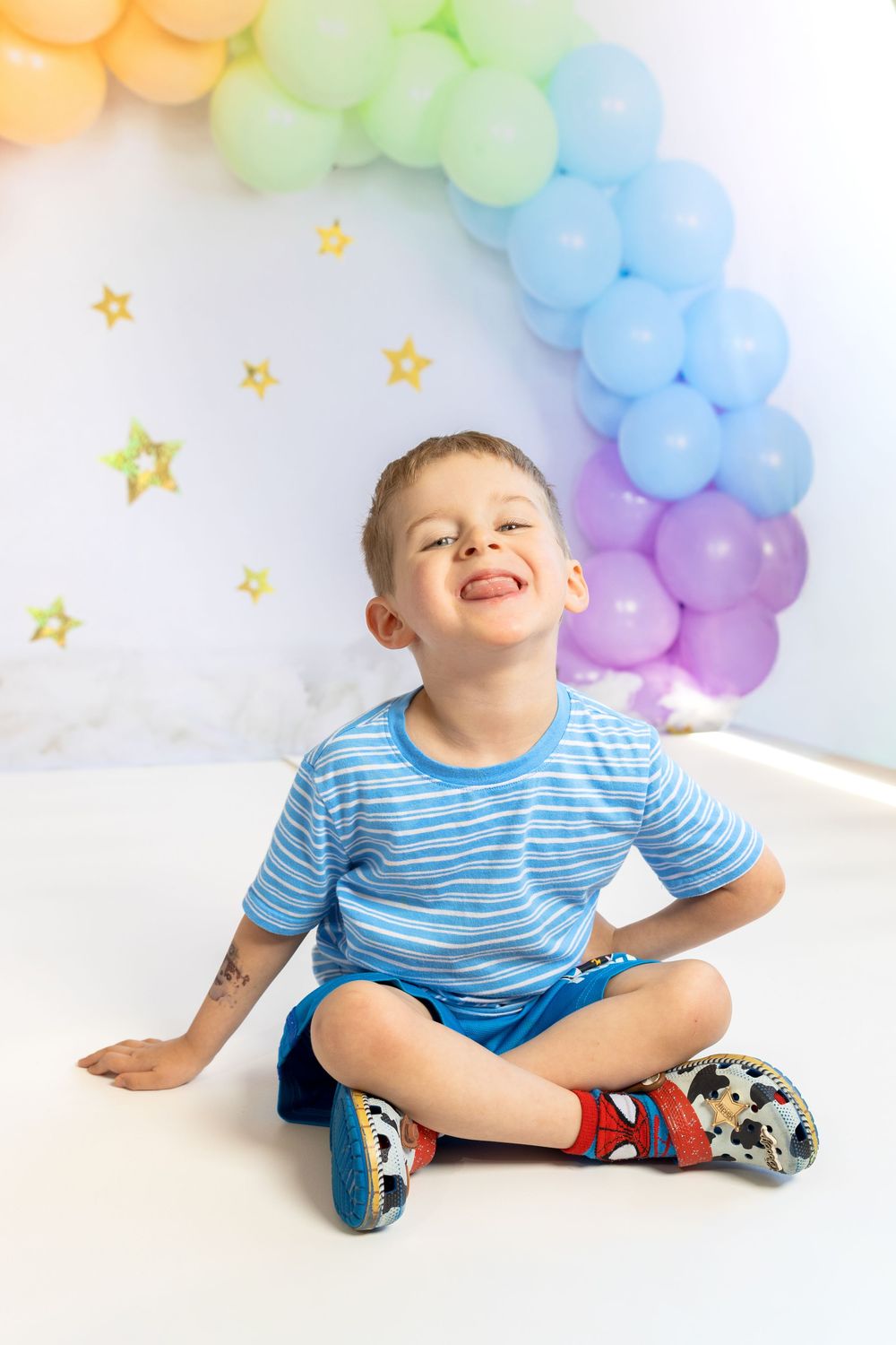 A cheerful boy sits cross-legged on white floor, sticking out tongue, with colorful rainbow balloon arch backdrop.