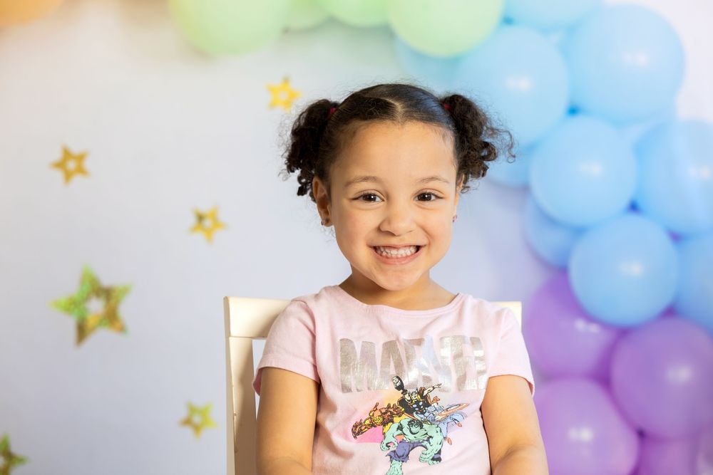 Smiling young girl with pigtails sits in front of colorful balloons and gold stars at a birthday celebration.