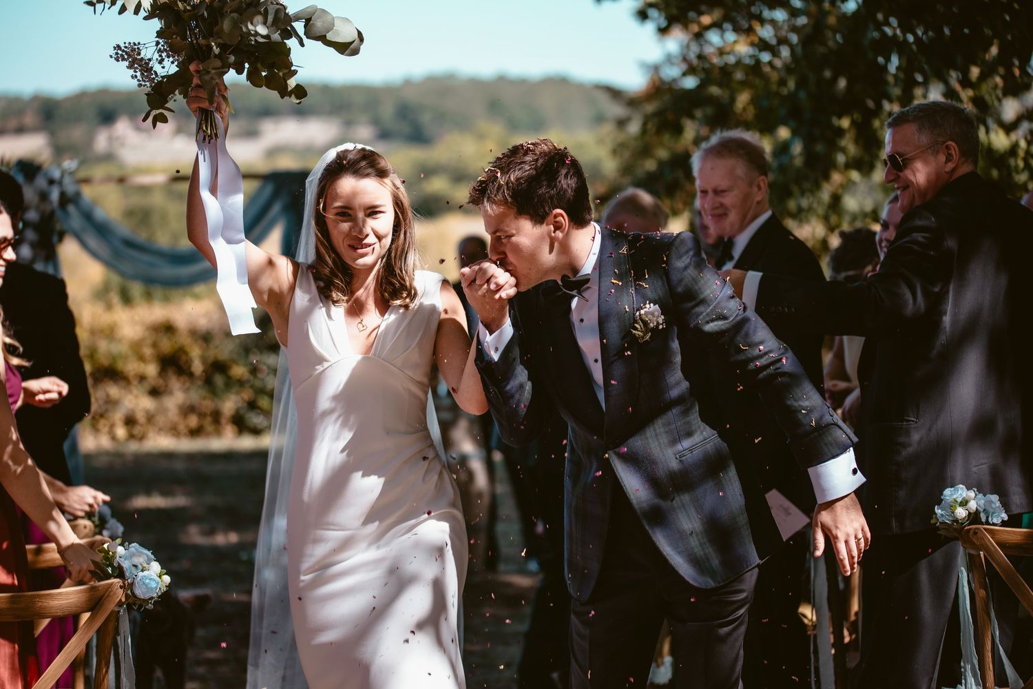 Beth raising her bouquet as Matt kisses her hand during their confetti exit.