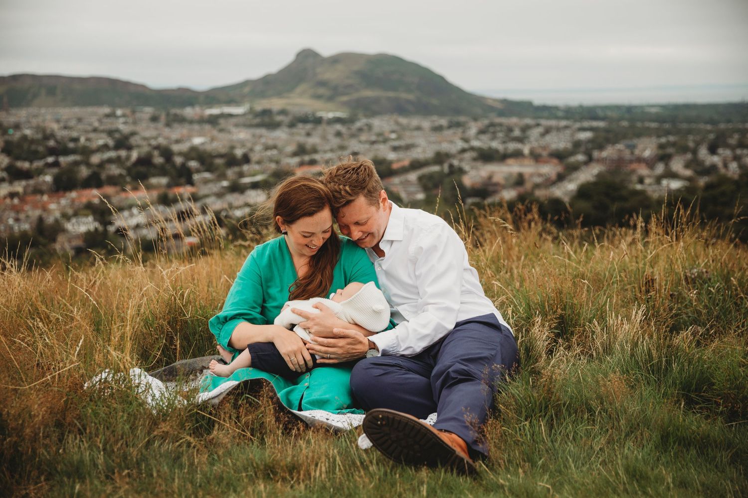 Couple sitting on grassy hilltop overlooking city view while holding newborn baby.
