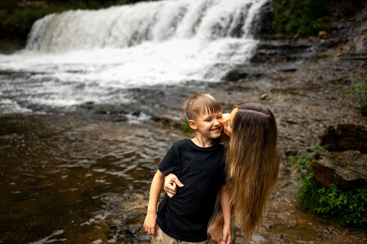 A mother kissing her young son near a scenic waterfall surrounded by rocks and lush greenery.