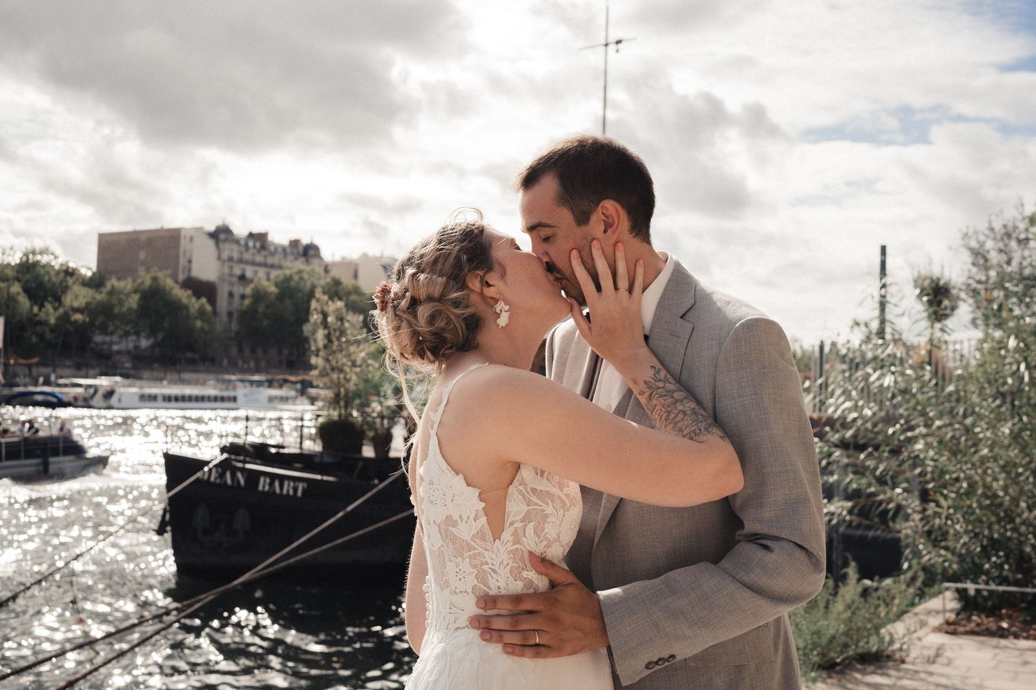 Newlywed couple sharing a romantic kiss in wedding attire by a scenic waterfront with boats and city skyline.