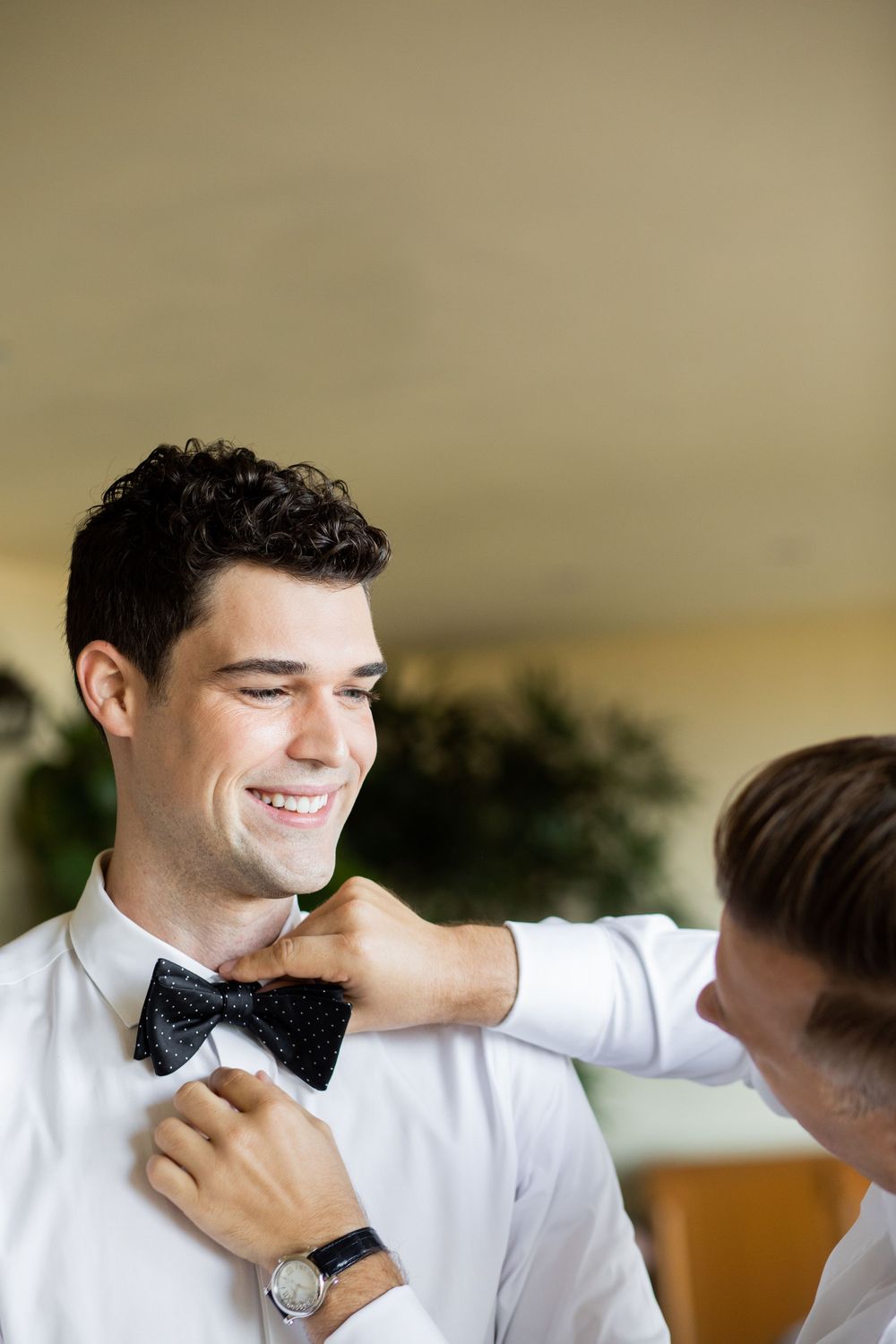 Smiling groom in white shirt getting black polka dot bow tie adjusted by groomsman before wedding ceremony.