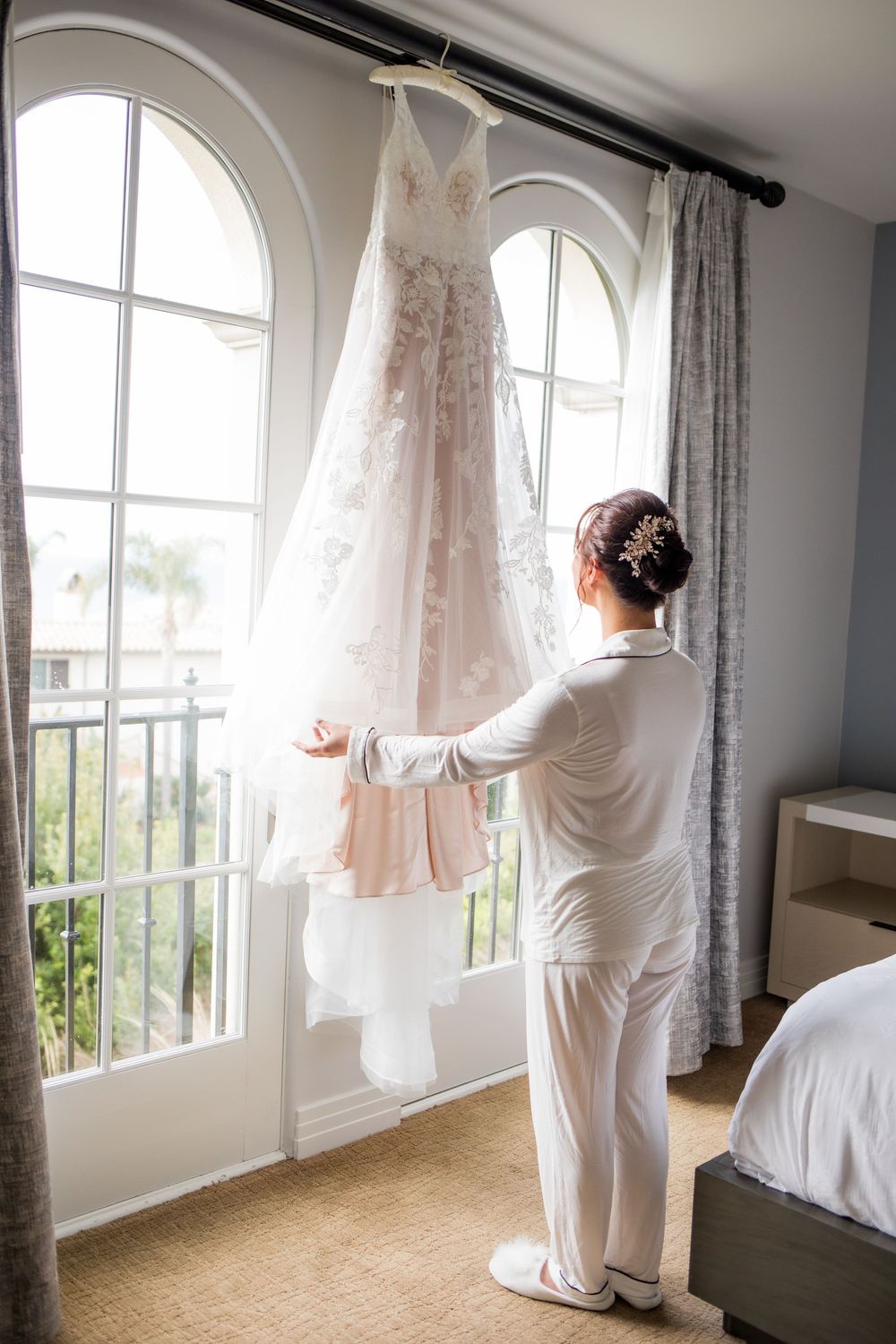 Bride in white robe admiring her lace wedding gown hanging by arched windows in a bright hotel room.