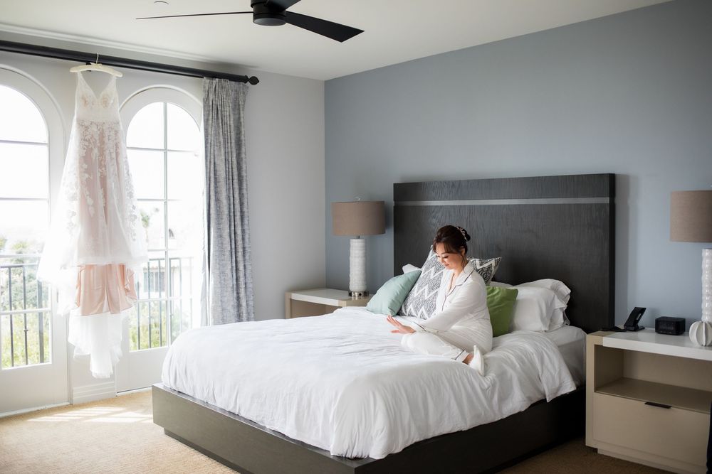 Bride sitting on white bed in blue bedroom with wedding dress hanging by arched window and ceiling fan above.