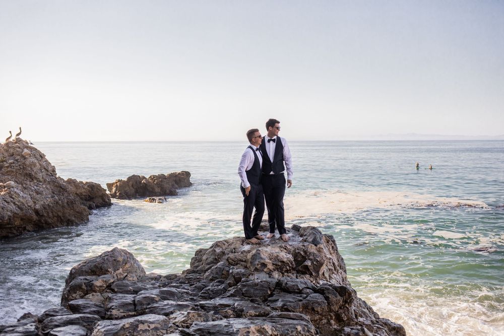 Two grooms in formal attire stand on coastal rocks with ocean waves and clear sky backdrop.