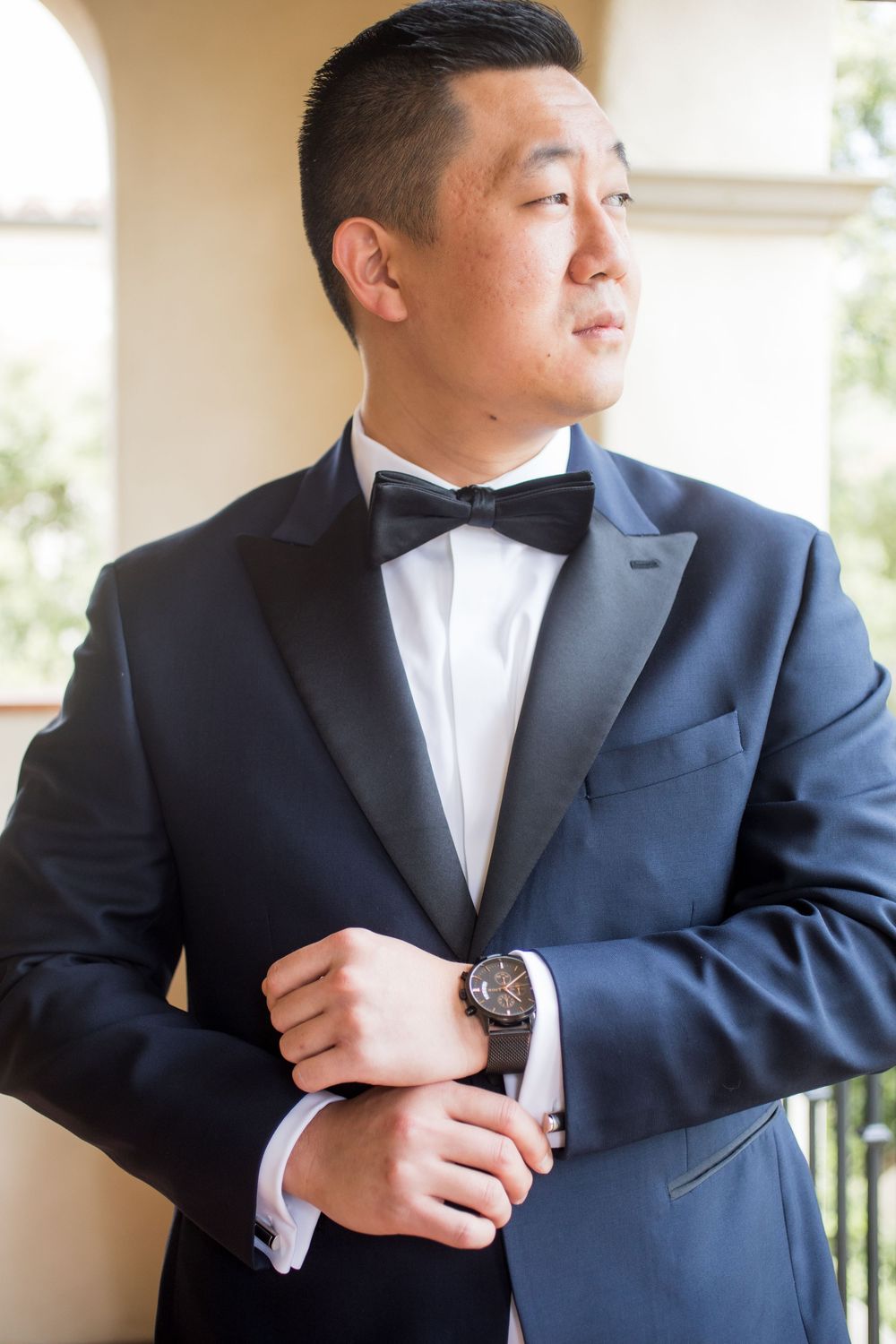 Groom in navy tuxedo with black bow tie adjusting his watch cufflinks before wedding ceremony.
