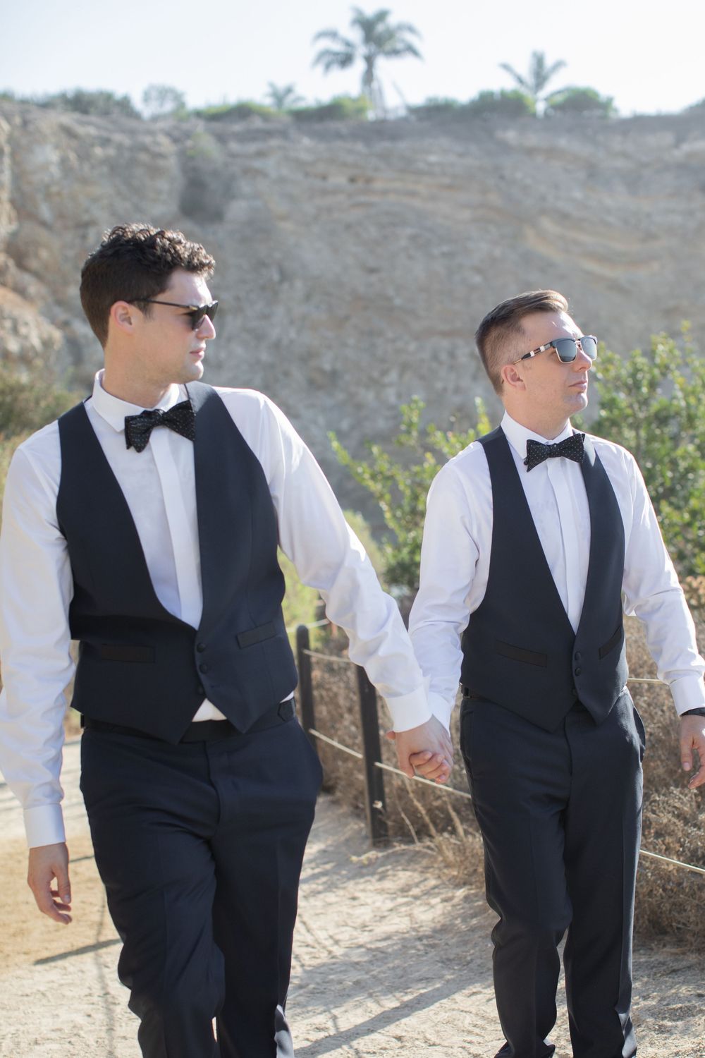 Two grooms in matching black vests and bow ties hold hands outdoors on a sunny day.
