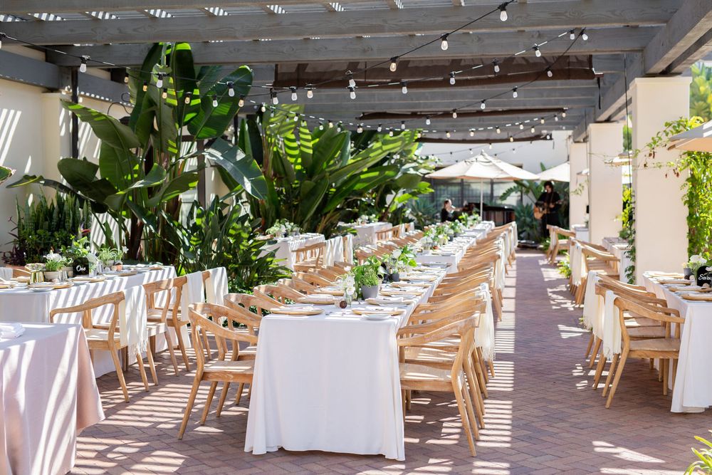 Elegant outdoor wedding reception with white tablecloths, wooden chairs, string lights, and lush tropical greenery under a pergola.