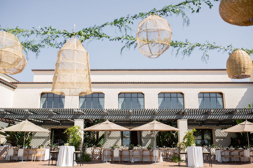 Elegant outdoor wedding reception with wicker pendant lights, greenery garlands, and draped tables under a pergola.