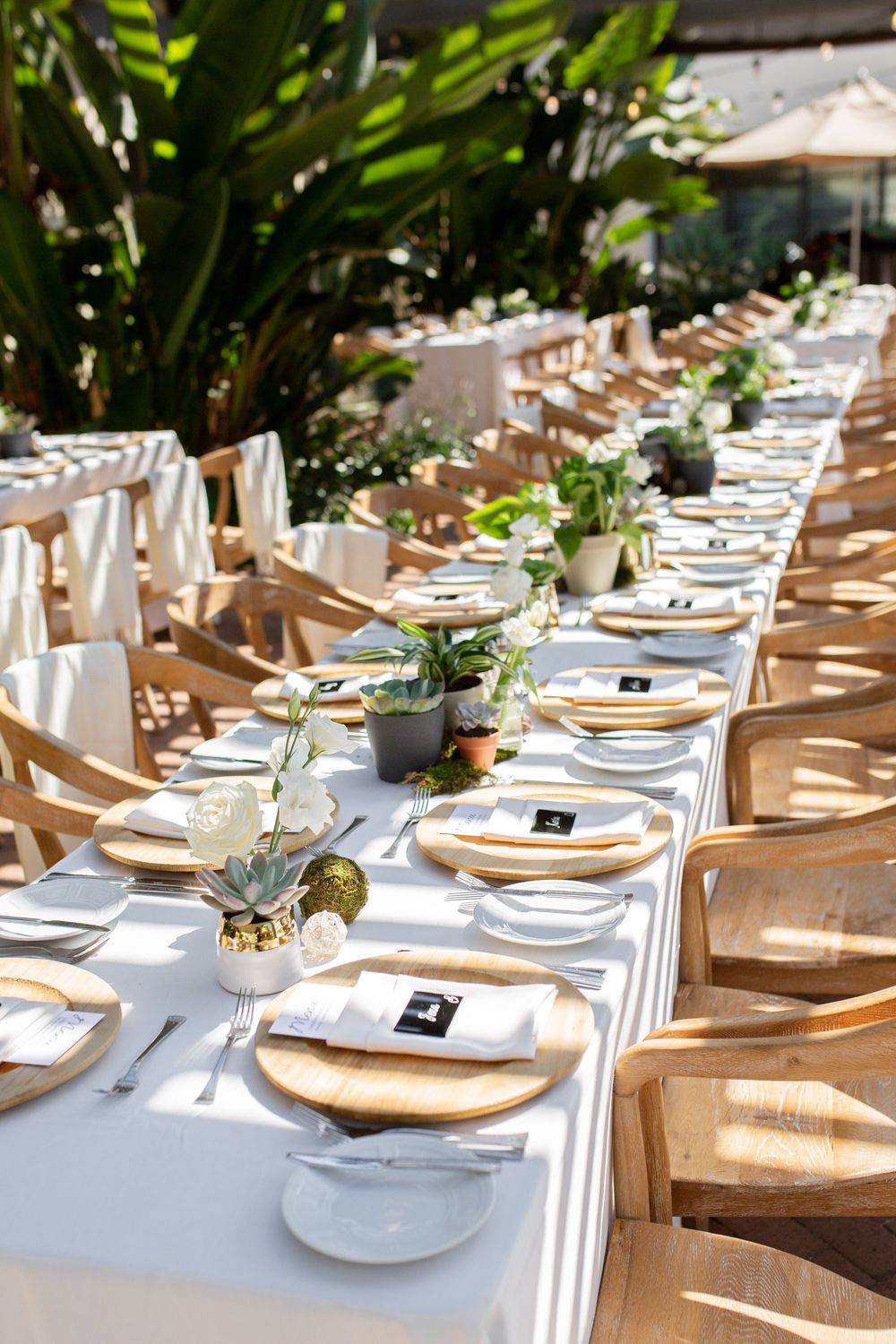 Elegant outdoor wedding reception with long white tables, wooden chargers, greenery centerpieces, and natural wood chairs.