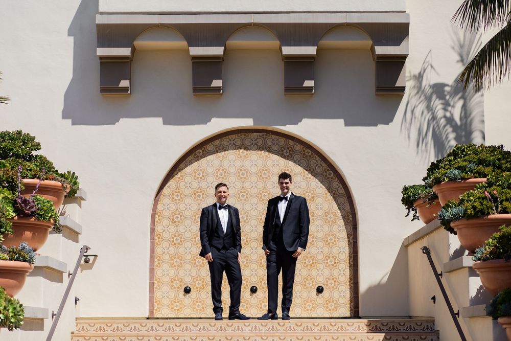 Two grooms in black tuxedos stand before an elegant arched doorway at an outdoor wedding venue.