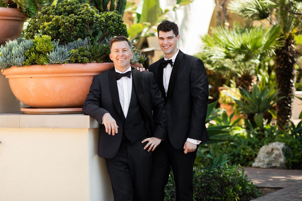 Two men in black tuxedos smiling together outdoors among lush tropical greenery at a formal event.
