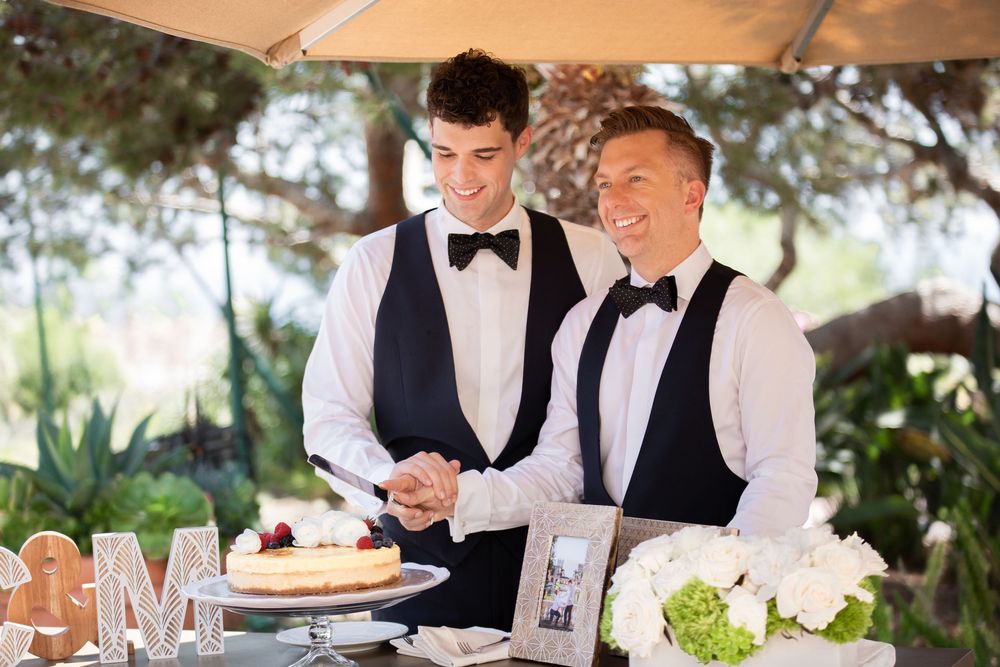 Two grooms in matching vests and bow ties cutting their wedding cake outdoors at an elegant reception.