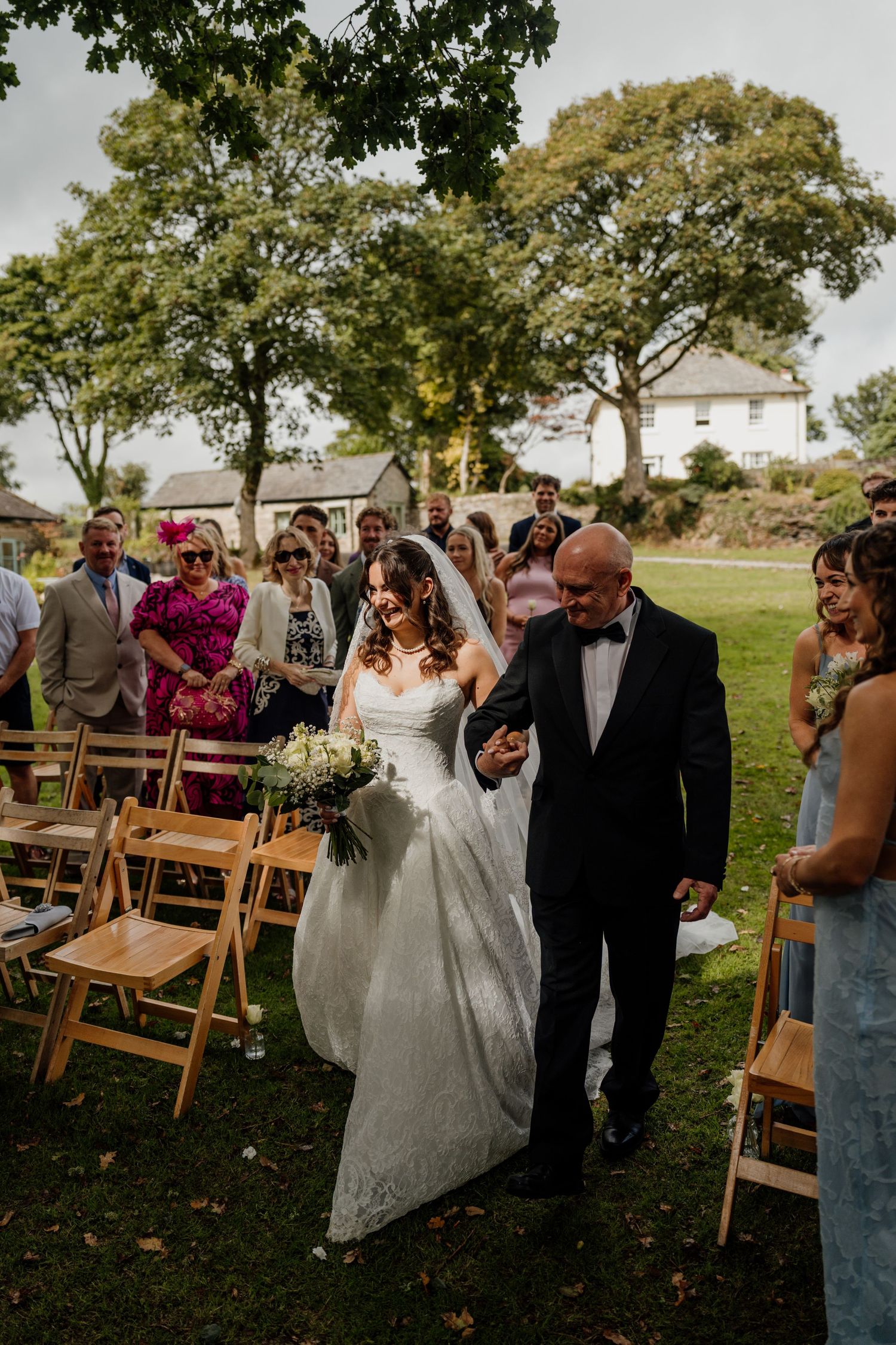 Bride walks down outdoor aisle with father at garden wedding ceremony at The Green wedding venue in Liskeard.