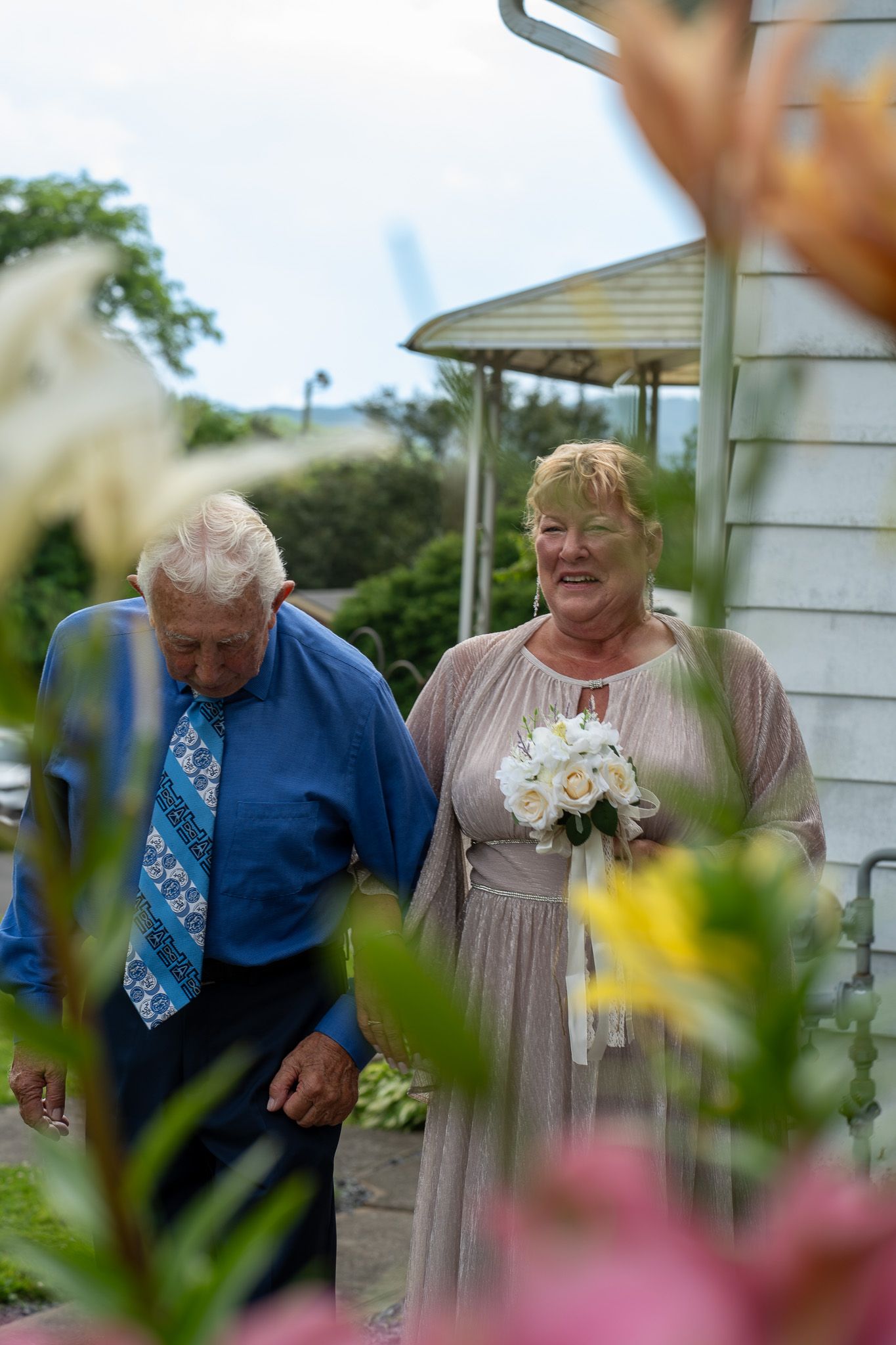 Elderly couple walking together outdoors, woman holding white bouquet in beige dress, man in blue shirt and tie.