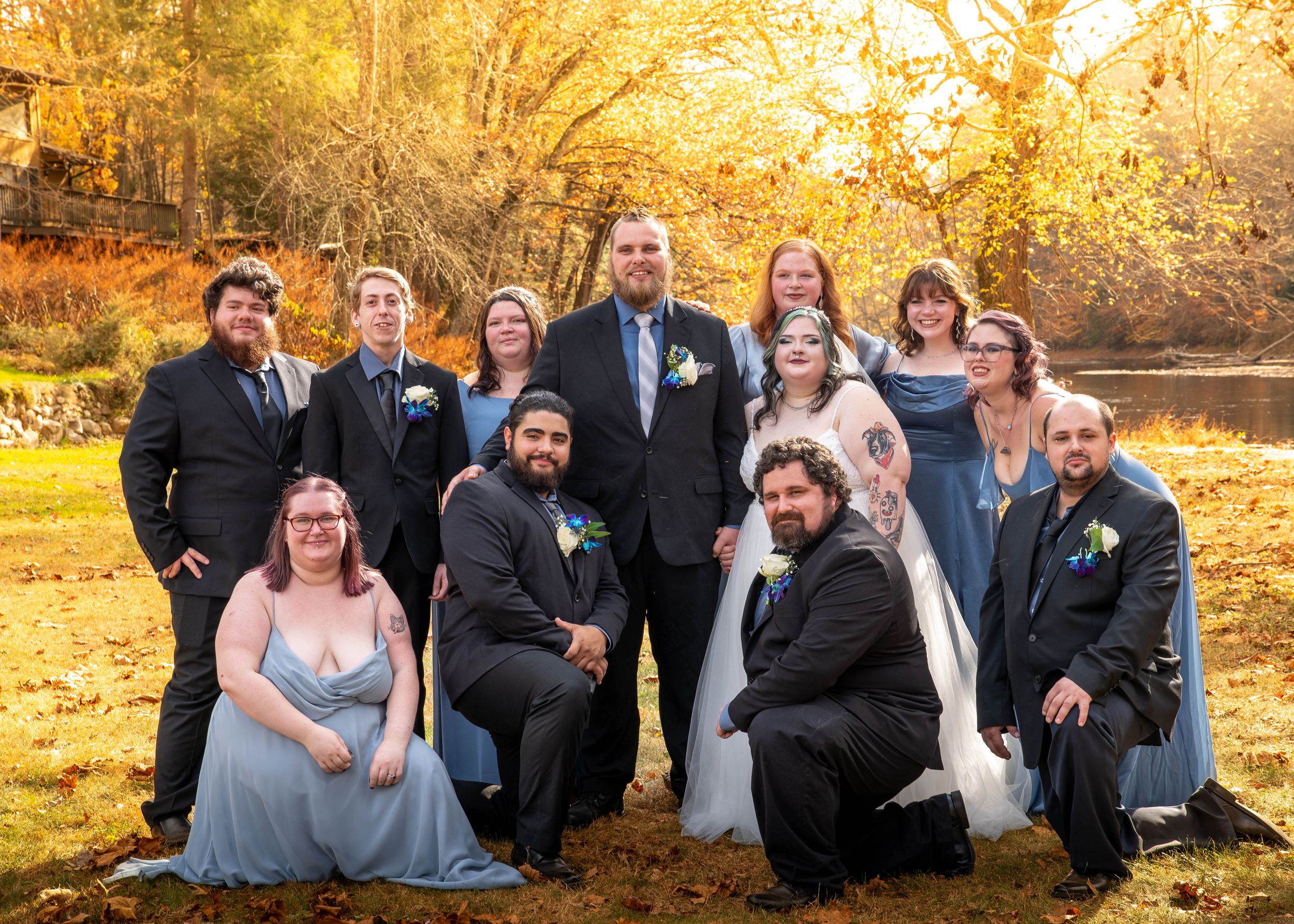 Wedding party poses outdoors in autumn, bride and groom centered, attendants in blue and black formal wear, fall foliage backdrop.