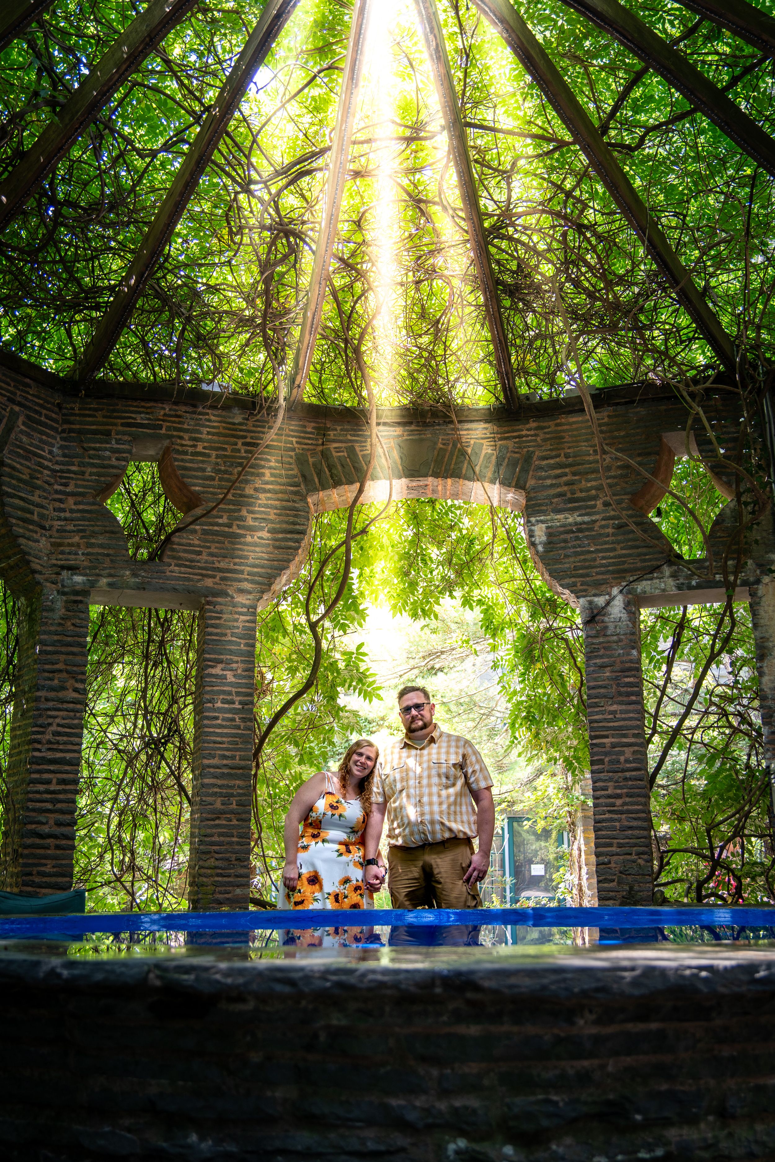 Couple holding hands inside a vine-covered brick gazebo with sunlight streaming through the leafy canopy above.