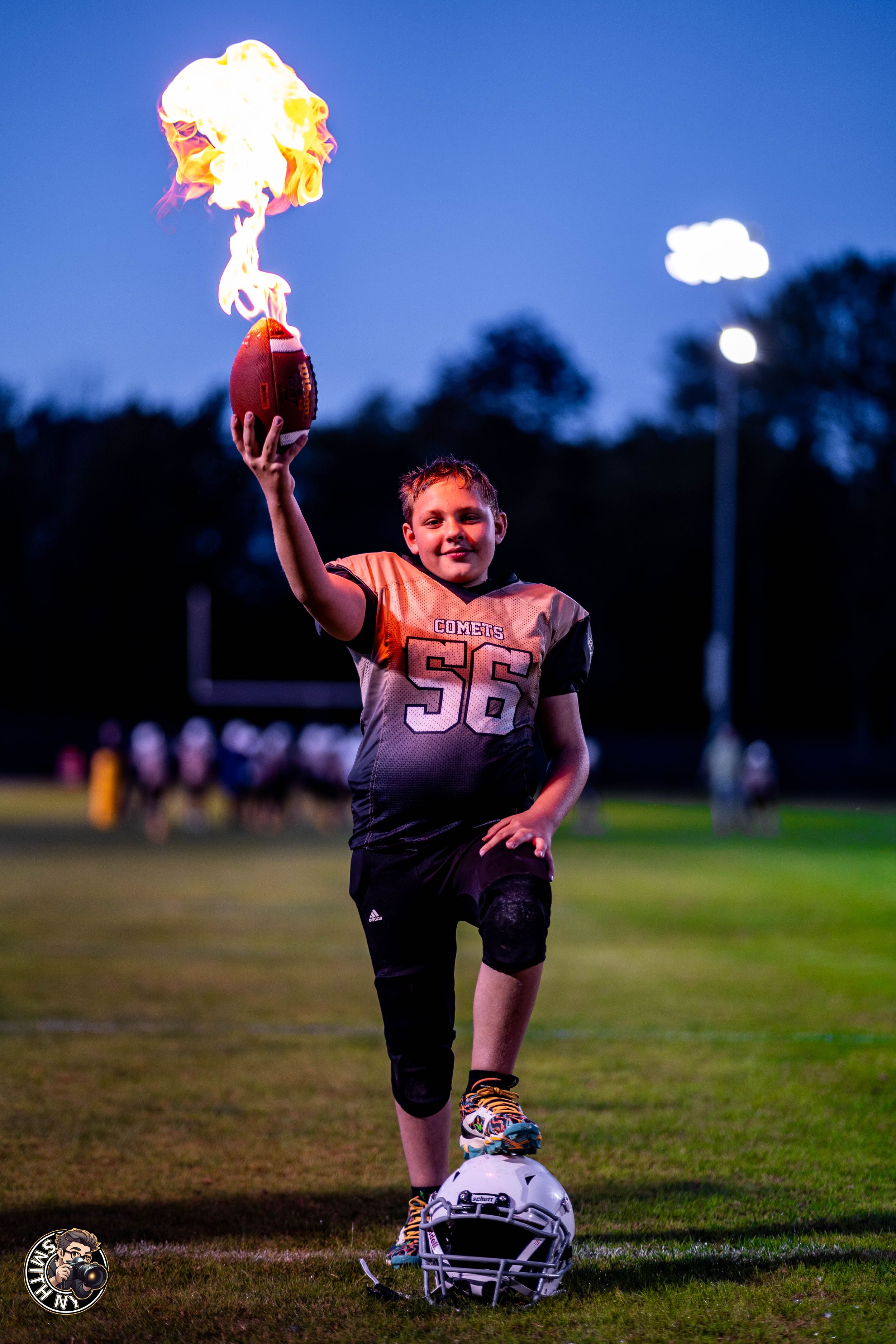 Youth football player #56 holds a flaming football aloft on a field at dusk, wearing a dark jersey and standing on a helmet.