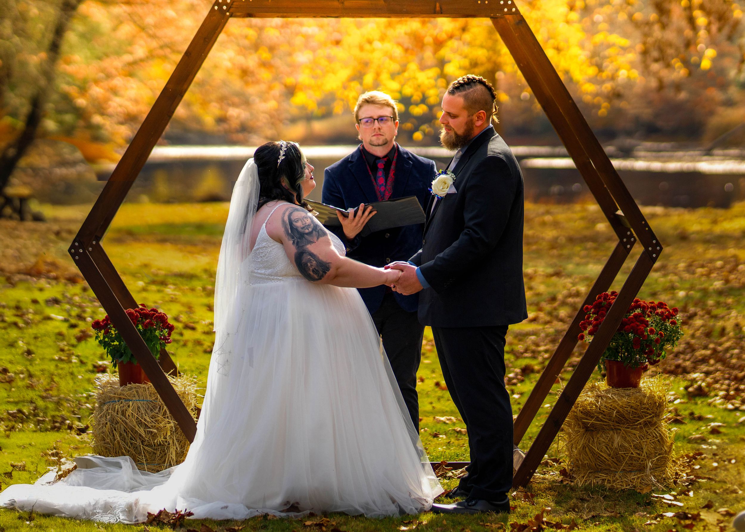 Couple exchanges vows at outdoor fall wedding ceremony under hexagonal arch with officiant, surrounded by autumn foliage.