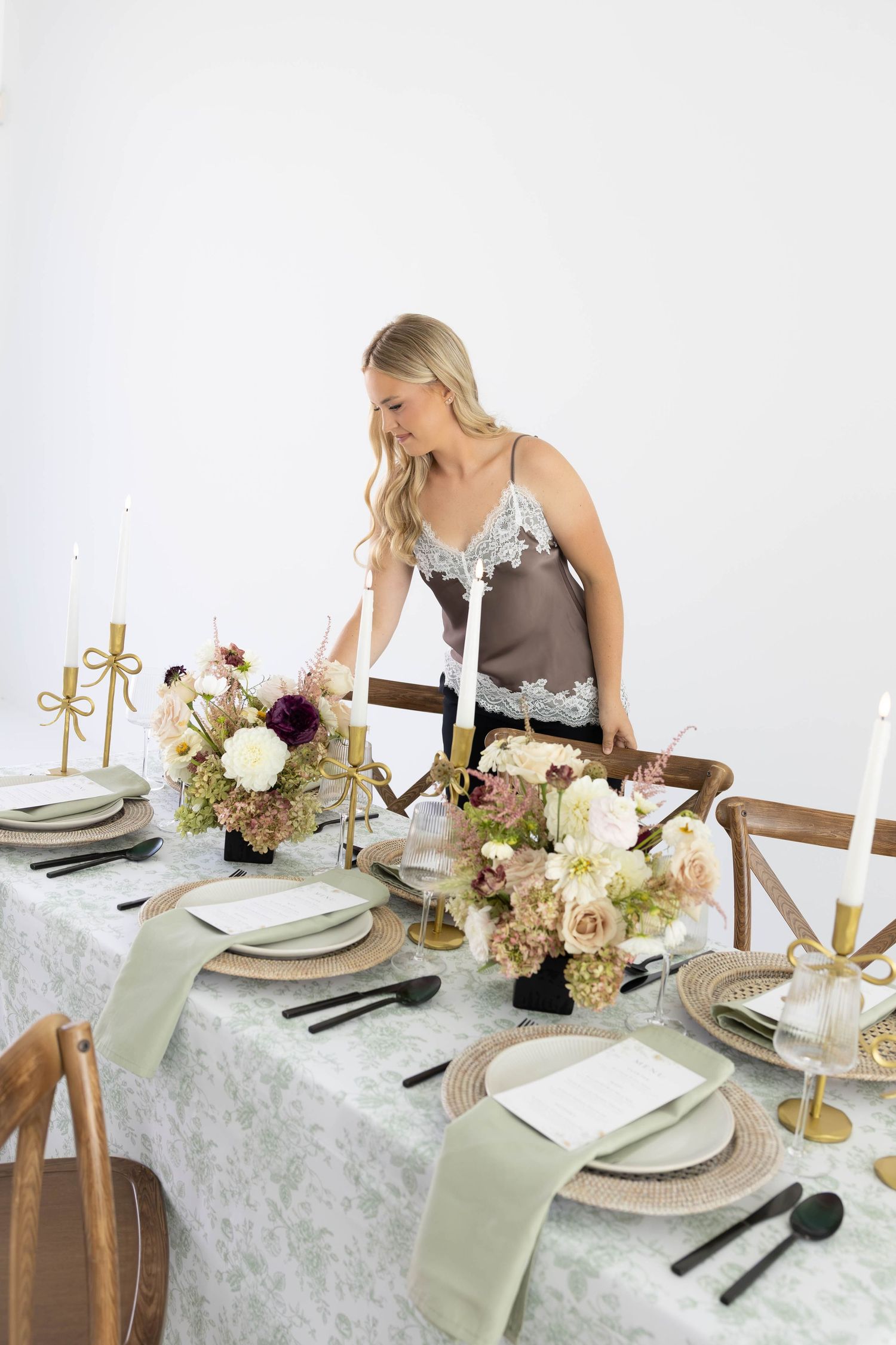 Blonde woman arranging elegant floral centerpiece on lace-covered reception table with gold candlesticks and black cutlery.