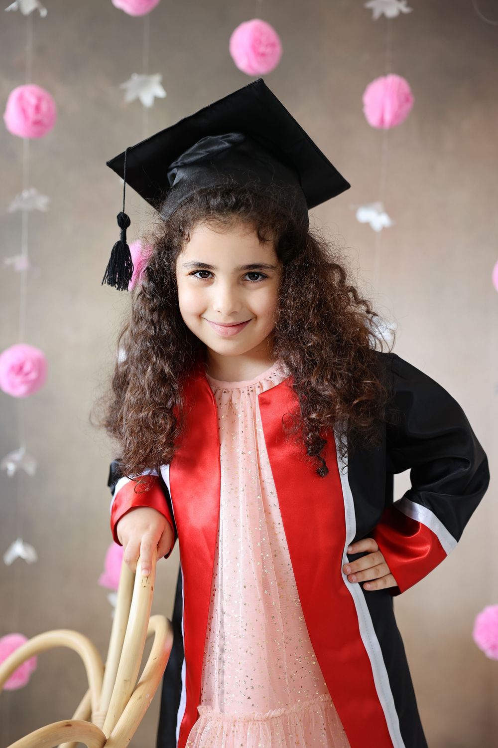 Young girl in graduation cap and gown smiling confidently, with pink decorations in background.