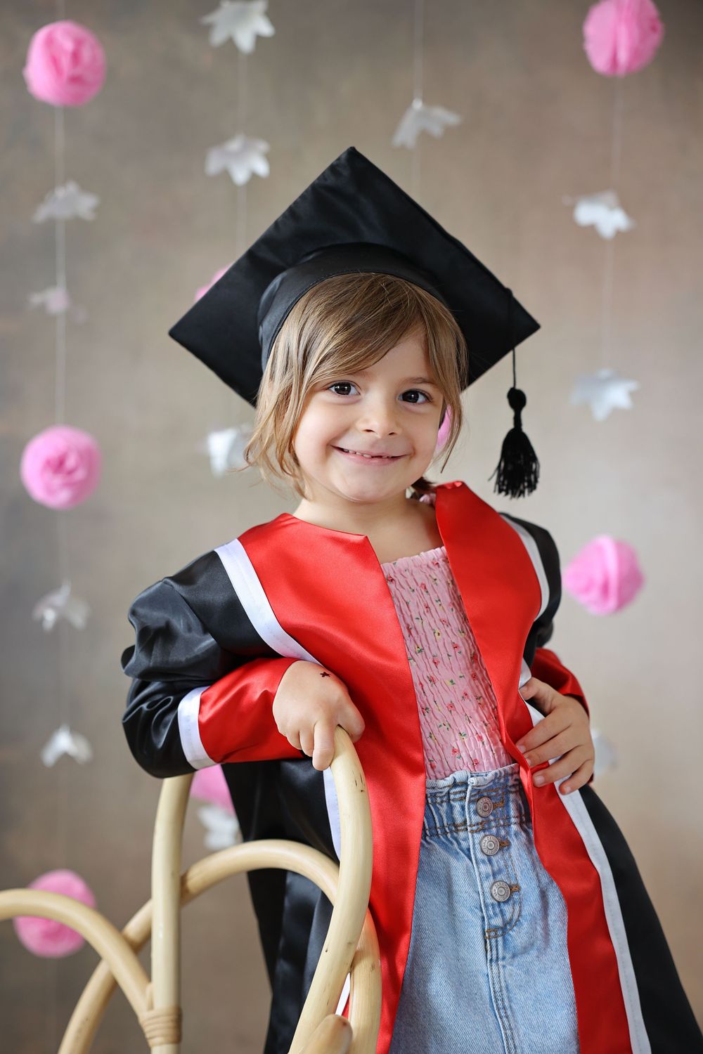 Smiling toddler girl wearing black graduation cap and red academic gown poses on wicker chair with pink decorations.
