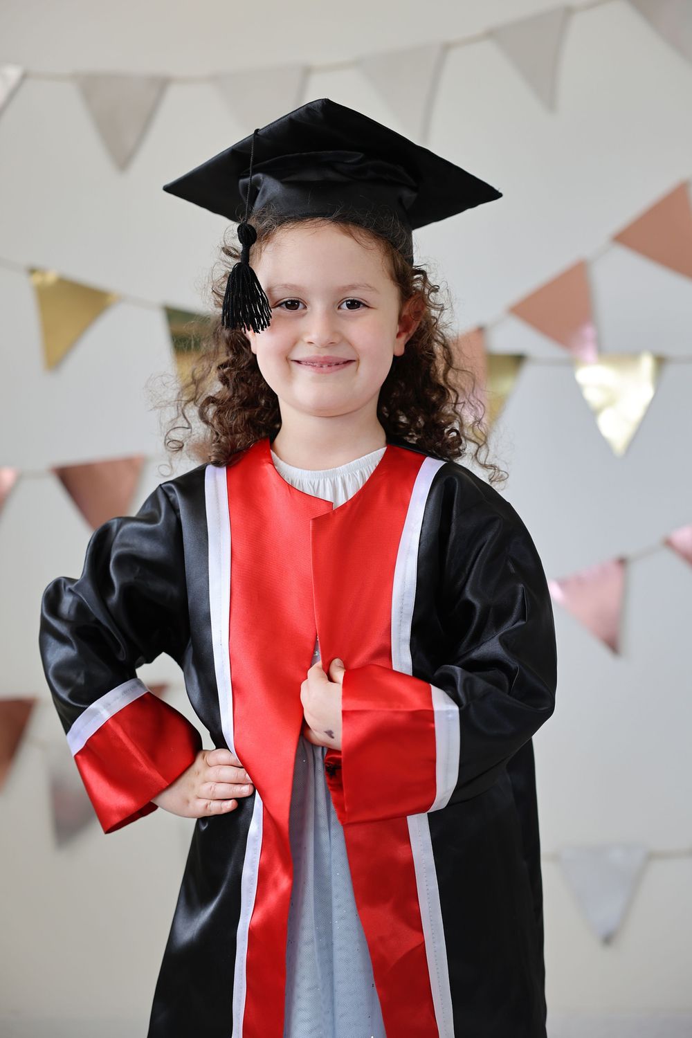 Young girl in black graduation gown, red sash, and mortarboard cap smiling at preschool graduation ceremony.
