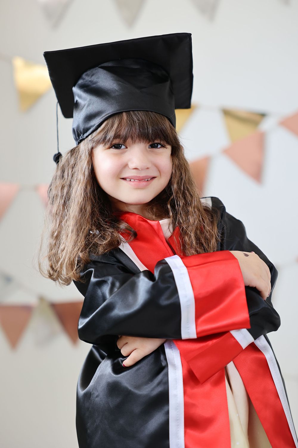 A smiling little girl in a black graduation cap and gown holding a diploma with a red sash at her graduation ceremony.