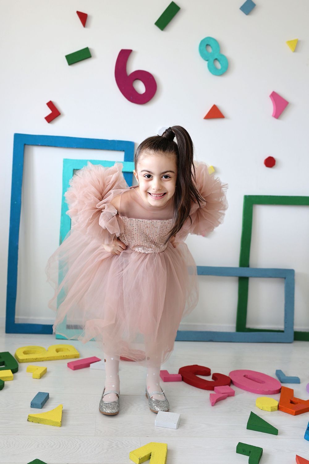A smiling girl in a pink tulle dress poses among colorful numbers and shapes in a bright, playful educational setting.