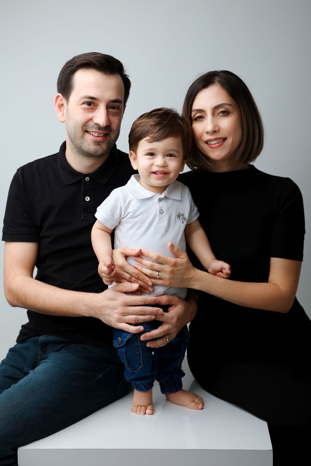 A smiling family of three poses for a studio portrait, with parents in black outfits holding their happy toddler boy.
