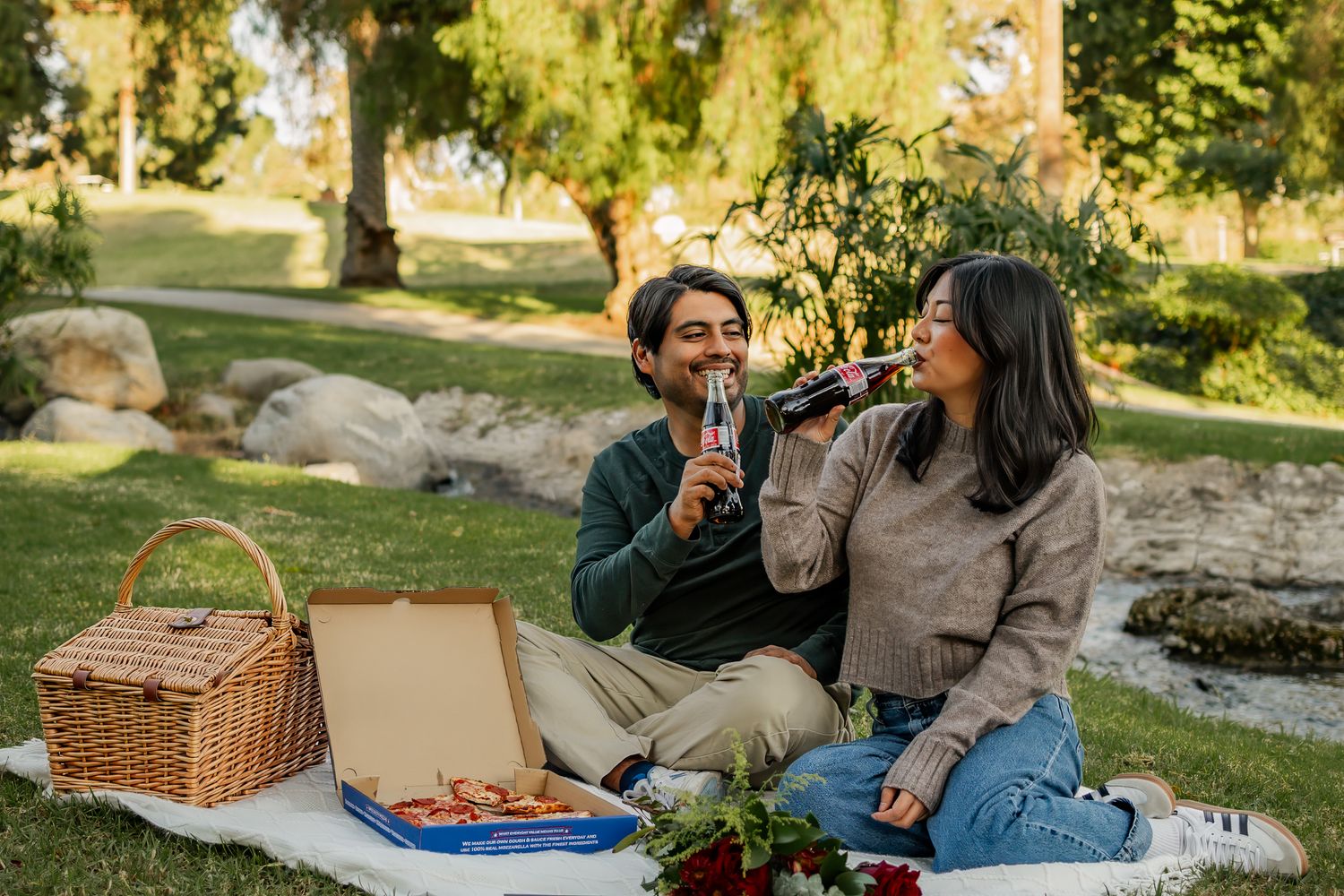Couple enjoying a romantic park picnic on a blanket with a wicker basket, wine, and a book on green grass.