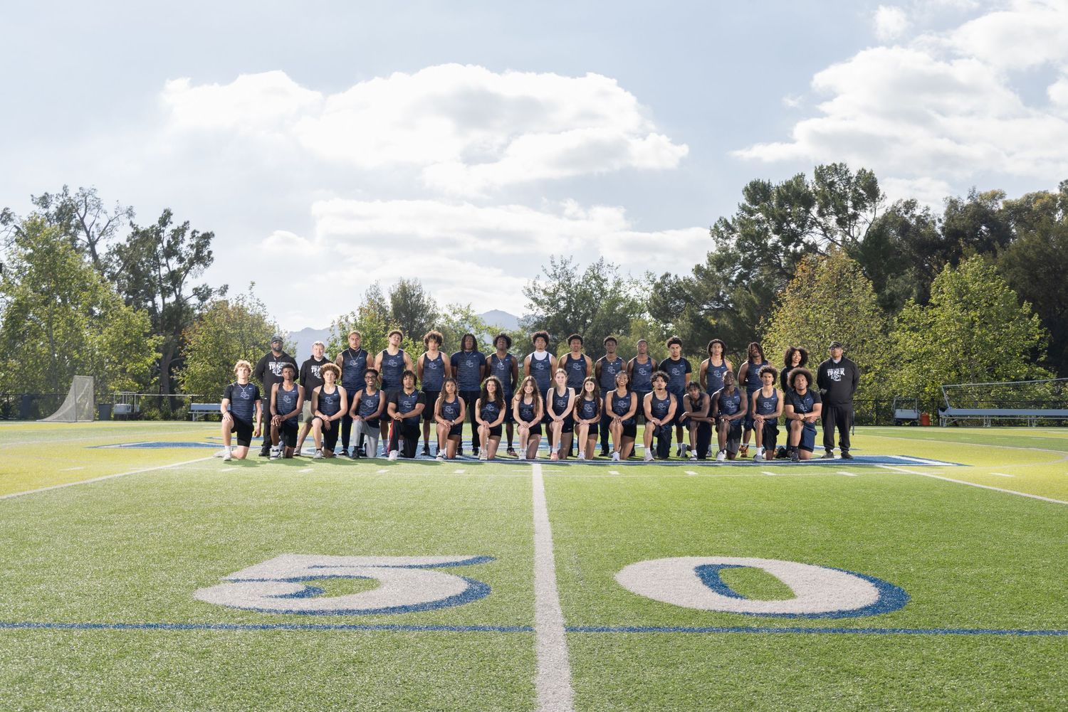 Sports team posing for group photo on football field at the 50-yard line, surrounded by green trees.