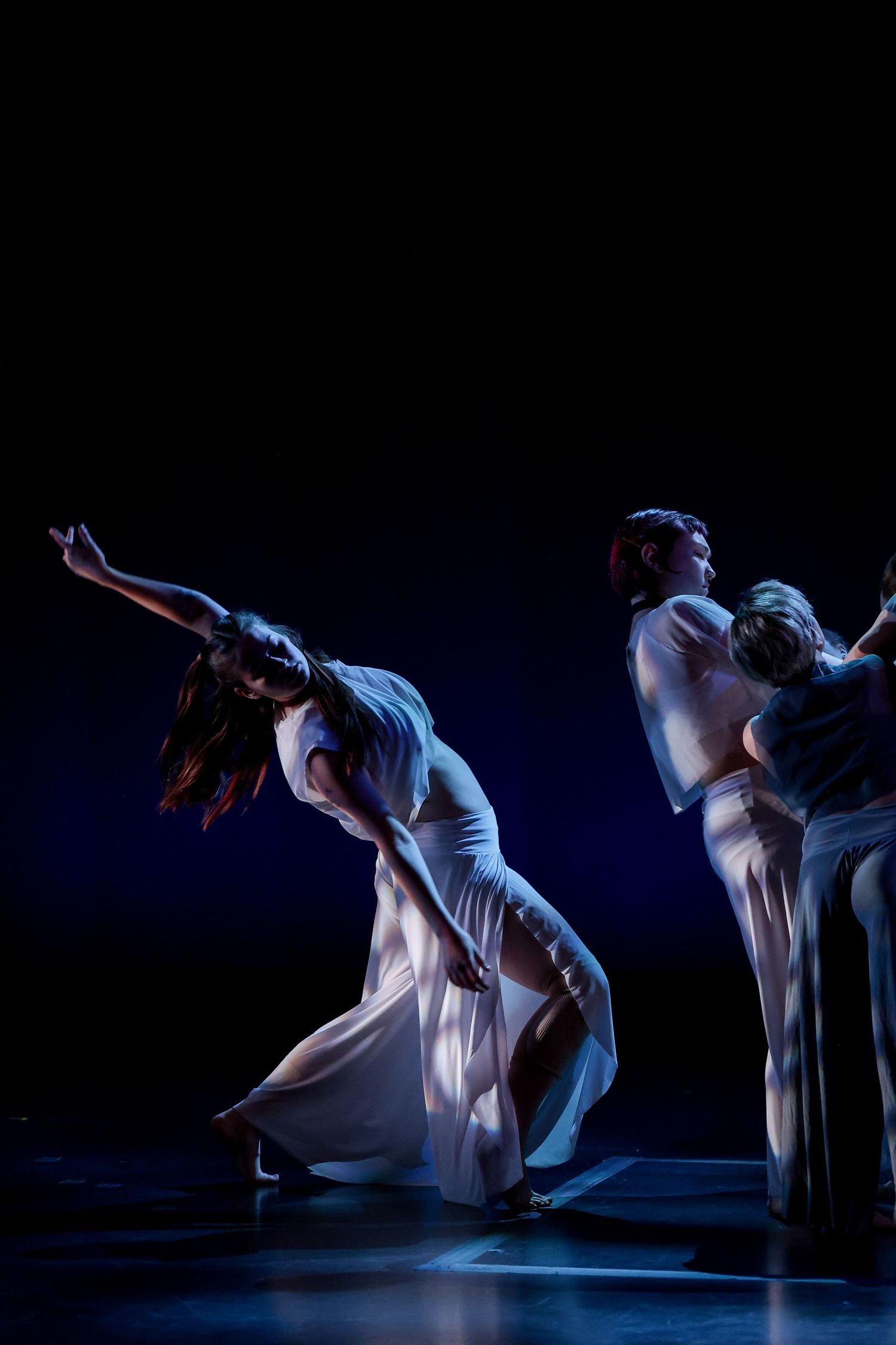 Dancers in white flowing gowns perform an expressive contemporary dance piece under dramatic blue stage lighting.