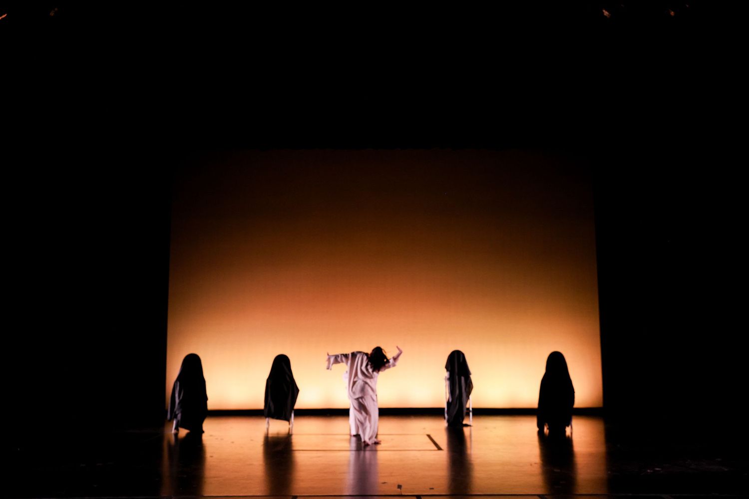 Performers in dark robes surround a central figure on a dimly lit stage with warm amber backdrop lighting.